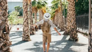 Traveler wearing a backpack and sun hat walking down a path lined with palm trees with arms stretched out.