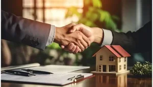 Real estate agent shaking hands with a client next to a model house and contract agreement.